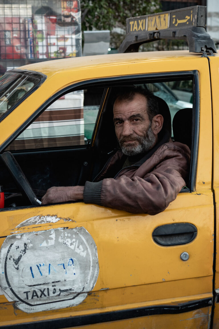 Yellow taxi parked under festive string lights and his driver looking at the camera in Damascus, Syria.