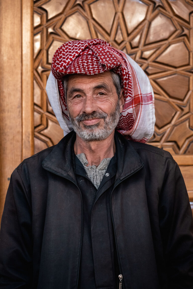 Elderly man wearing a red keffiyeh smiling in front of a traditional Syrian backdrop.