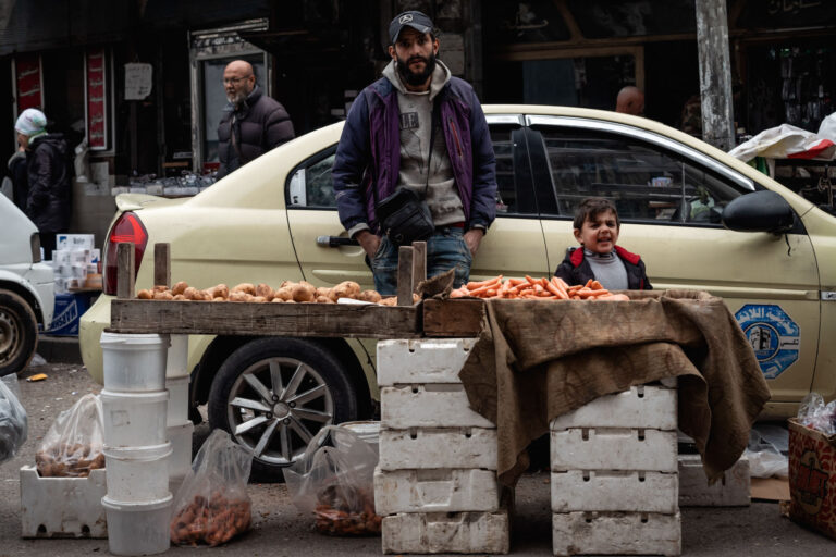 A young man in a jacket and a child selling oranges in the streets of a market in Latakia.