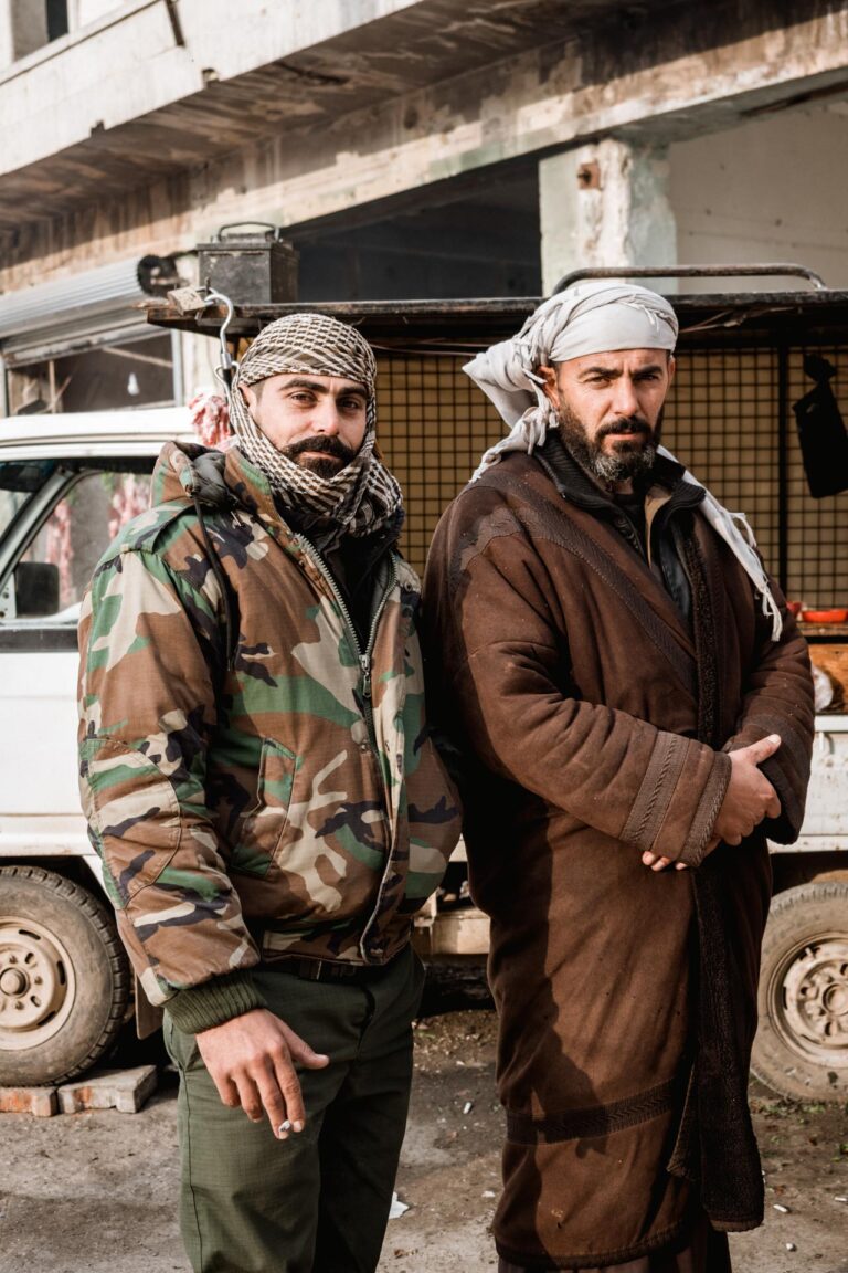 Two men in military clothing posing in front of a ruined building in Saraqib, Syria.