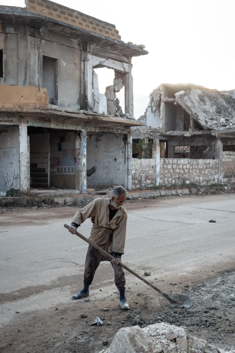 An elderly man working to rebuild his city, standing across a war-ravaged street in Saraqib, Syria.