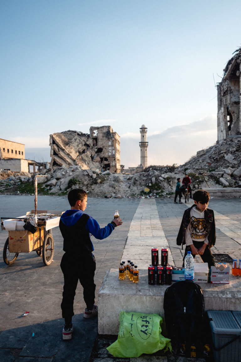 Kids selling stuff in the street, and behind remains of a minaret in a devastated landscape in Aleppo, Syria.