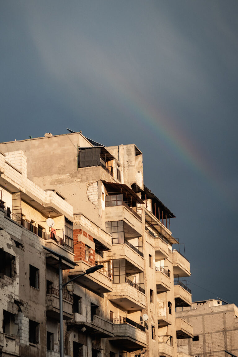 A war-torn urban landscape with partially collapsed buildings and a rainbow above, in Homs, Syria.