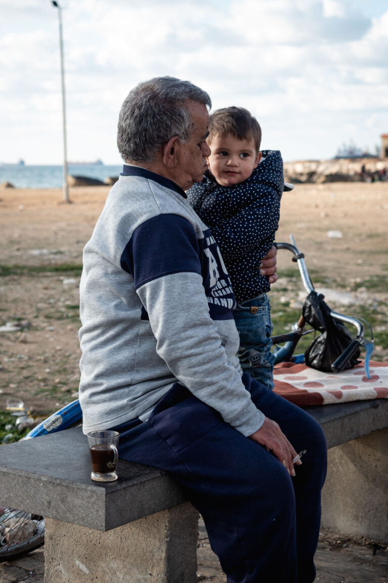 A grandfather and child sit on a bench, sharing a moment in Tartus, Syria.