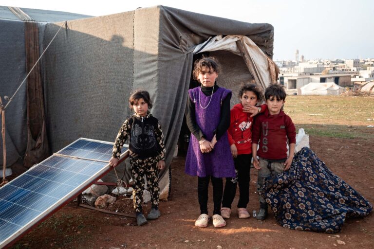 Four children stand near a tent in Saraqib, symbolizing Syria's humanitarian crisis.