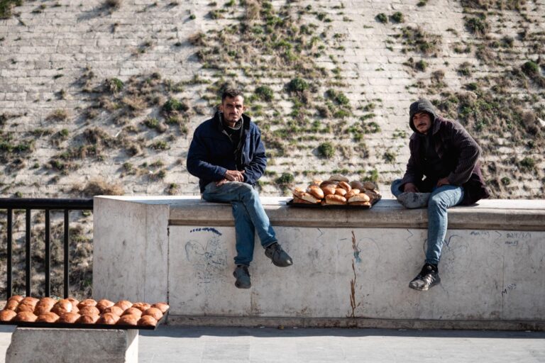 Two men sit by the citadel of Aleppo, Syria, selling fruits beside a historic site.