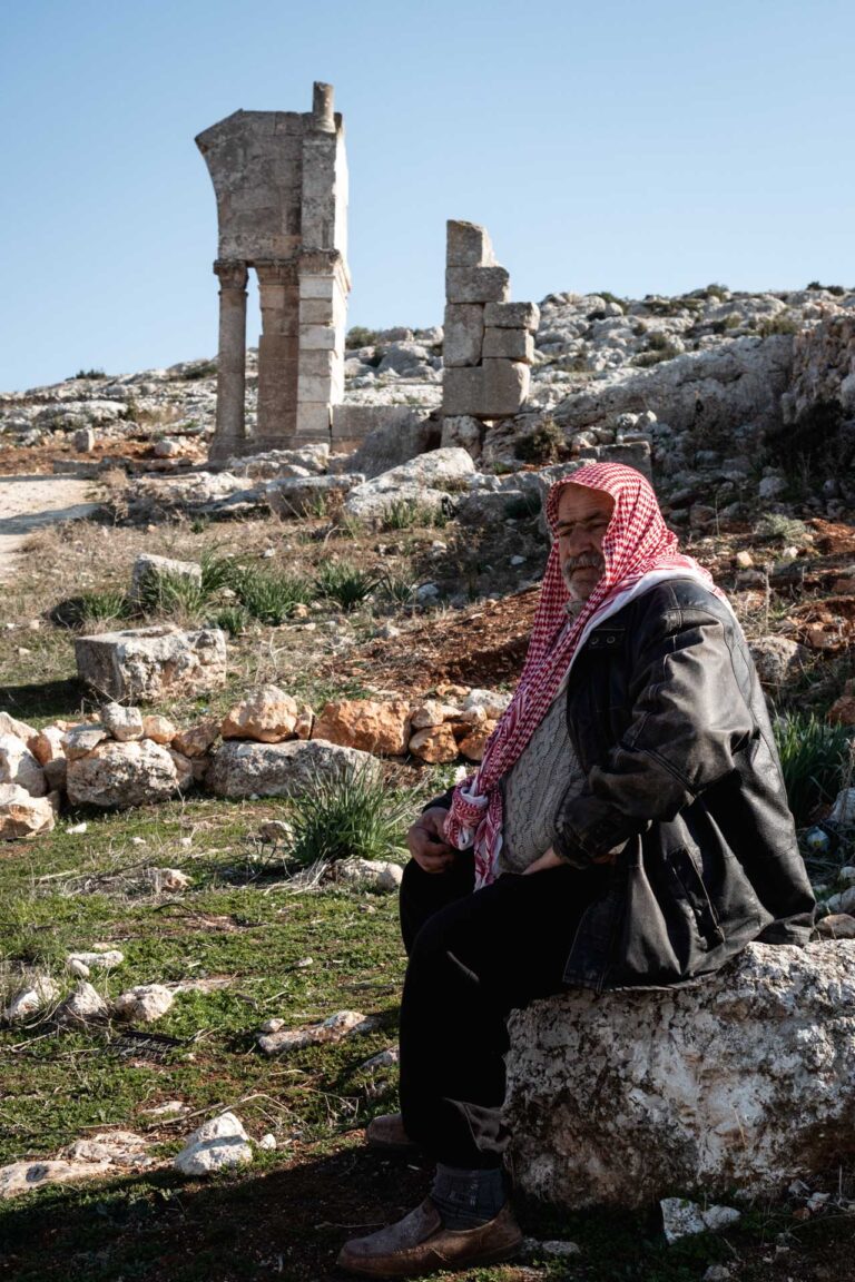 A man in a red scarf sits near ancient ruins in Saint Simeon, Syria.