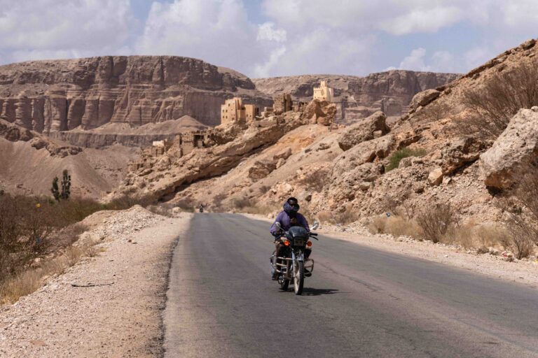 A person riding a motorcycle down a paved road through a rugged desert canyon.
