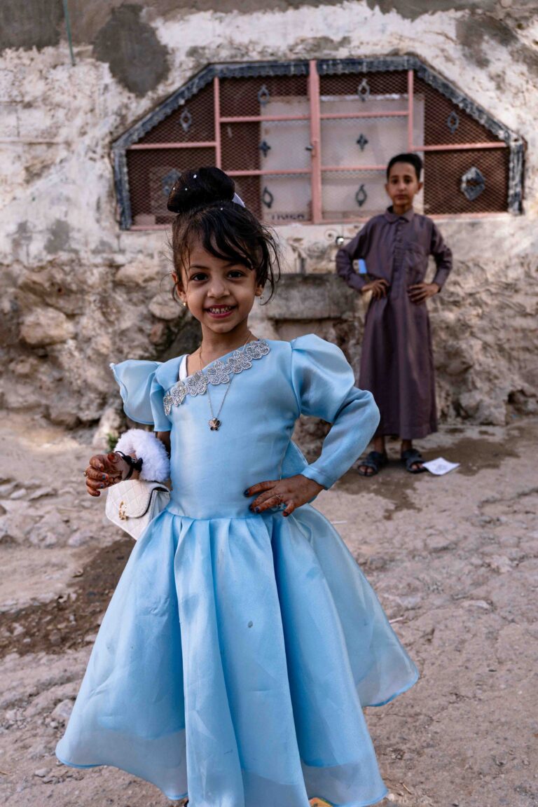 A young girl in a blue dress smiling in the village of Al Ribat.