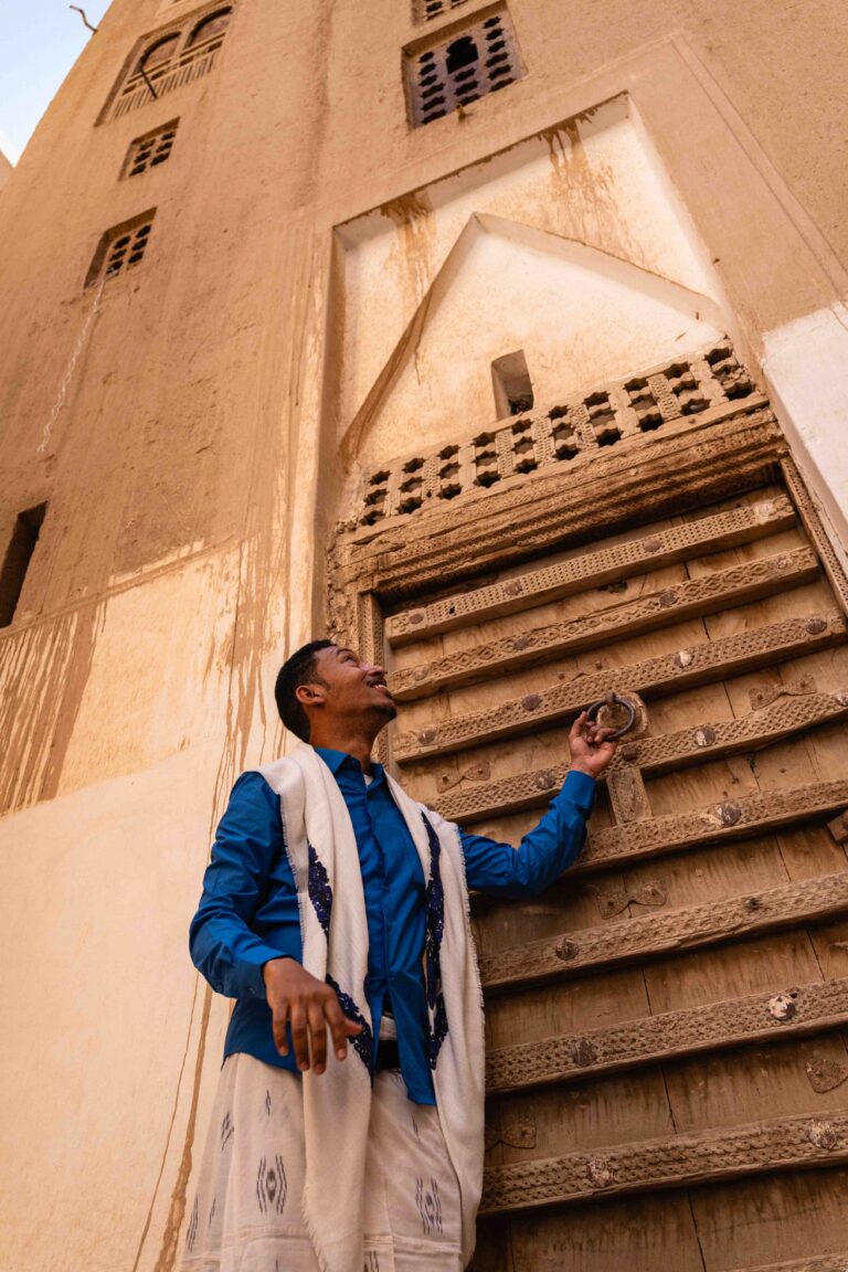 A man in traditional dress stands before a towering mud-brick building in Shibam.
