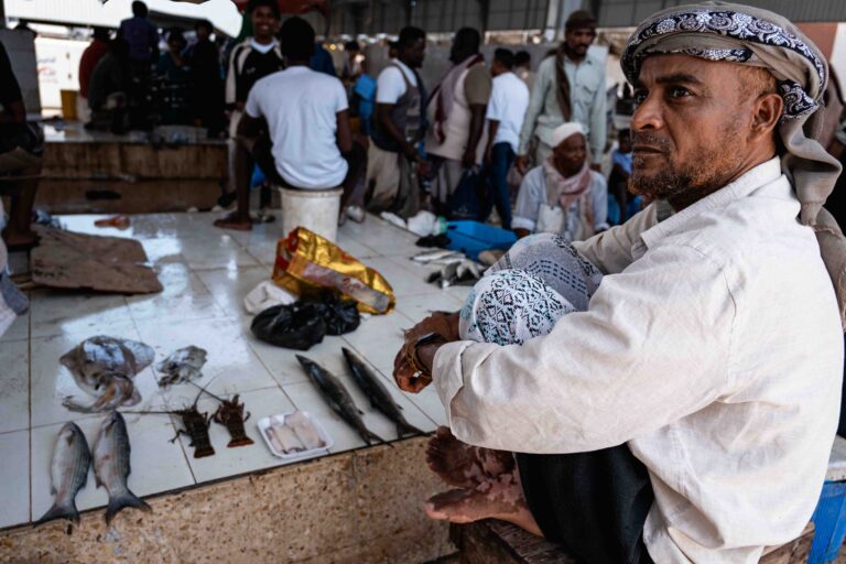 A man sitting in a fish market in Mukallah, Yemen, surrounded by local goods.
