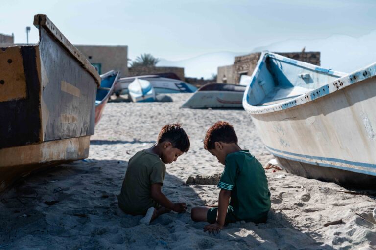 Two young boys playing in the sand next to fishing boats in Qalansiya.