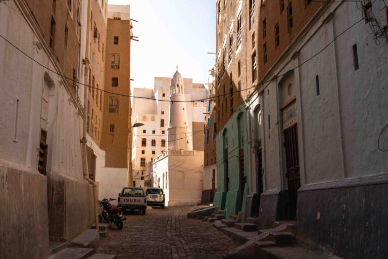 A narrow, sunlit street between the historic high-rise mud buildings of Shibam, Yemen.