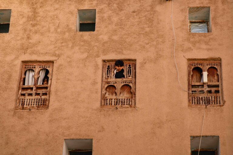 Close-up of ornate wooden windows with kids inside on a traditional mud-brick building in Shibam.