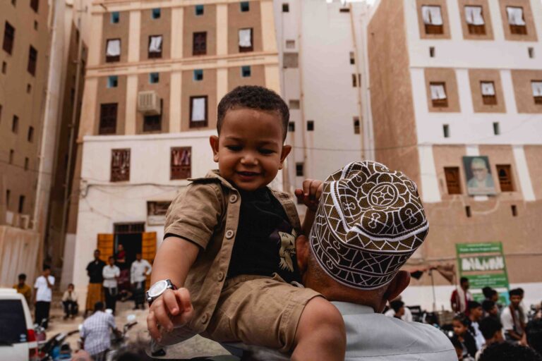 A young boy smiling while being carried through a crowded street in Shibam.